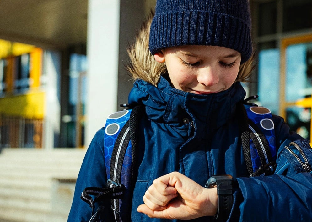 Dítě v zimním oblečení a čepici s batohem na zádech se usmívá a dívá se na dětské chytré hodinky